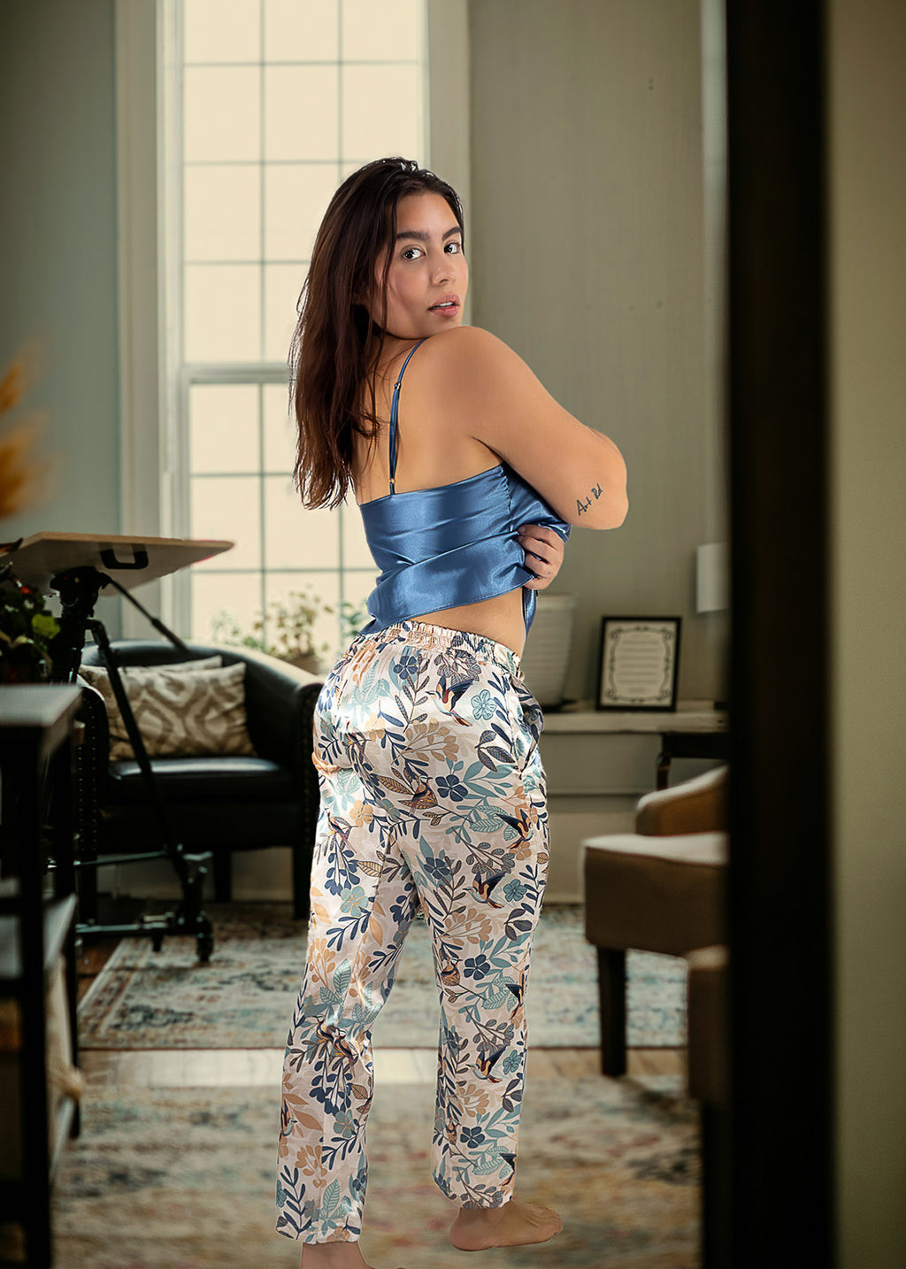 Woman in a blue top and patterned pants standing in a room with furniture and decor.